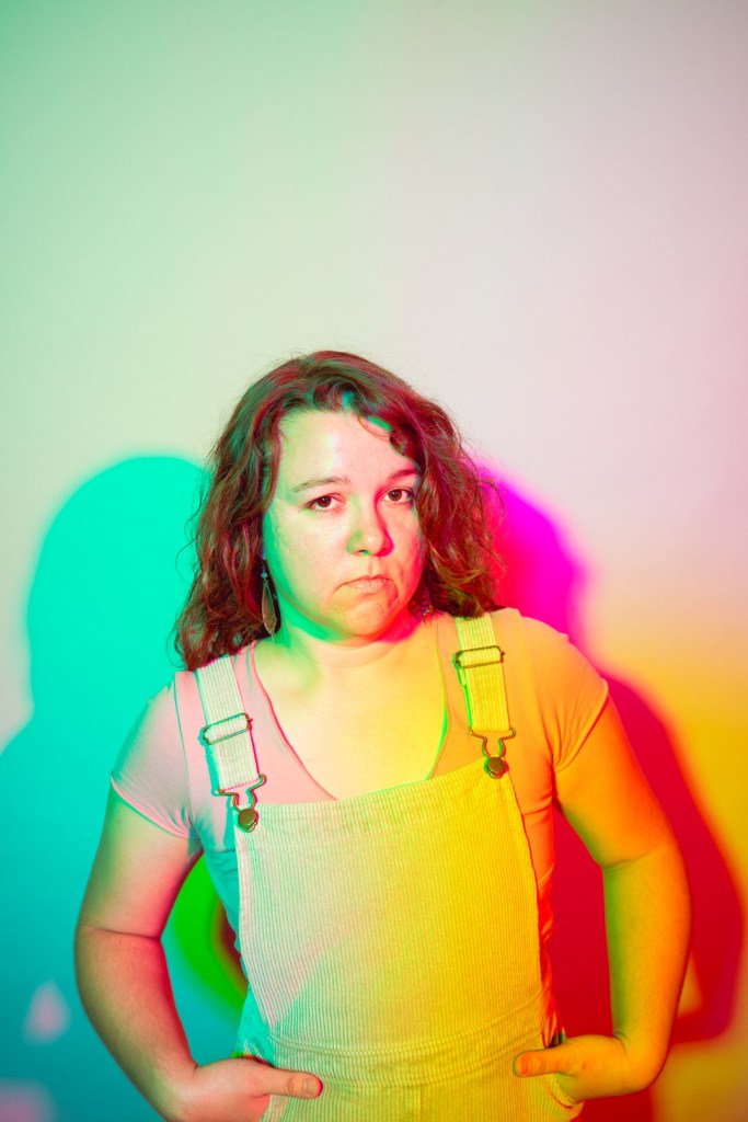 Meghan in overalls giving the camera a skeptical look under pink and teal light